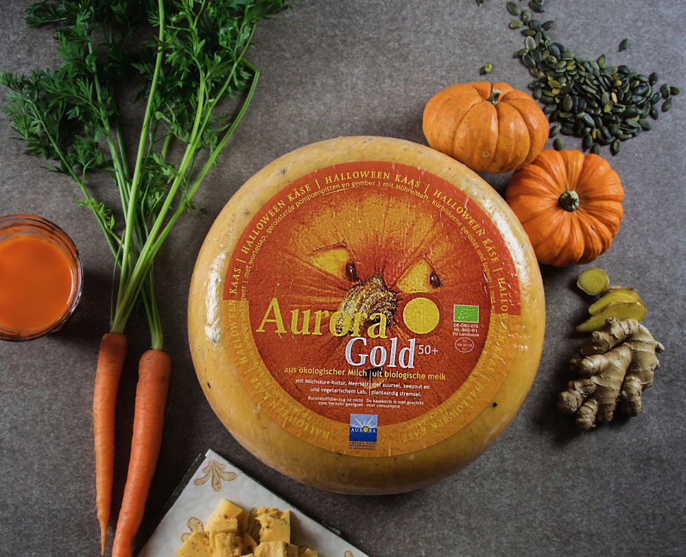 Runder Laib Aurora Gold Halloween-Käse mit Etikett, umgeben von Karotten, Kürbissen, Kürbiskernen, einem Glas Möhrensaft und einem Stück Ingwer