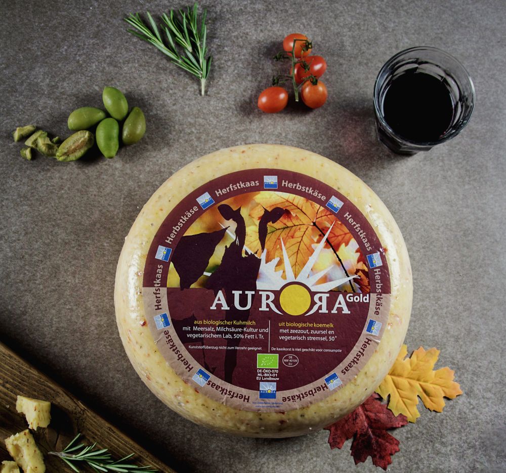 Runder Laib Aurora Gold Herbstkäse mit Etikett, umgeben von Rosmarinzweigen, grünen Oliven, Kirschtomaten, einem Glas dunkler Flüssigkeit und Herbstblättern.