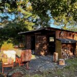 Im Hintergrund stehende Hütte aus Bretter, im Vordergrund stehen zwei rote Schaukelstühle mit Schaffellen. Neben und über der Hütte befinden sich Äste mit grünen Blättern.