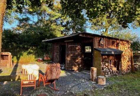 Im Hintergrund stehende Hütte aus Bretter, im Vordergrund stehen zwei rote Schaukelstühle mit Schaffellen. Neben und über der Hütte befinden sich Äste mit grünen Blättern.