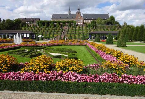 Im Hintergrund das Kloster Kamp, im Vordergrund ein Garten mit leuchtenden pinken und gelben Blumen an einer gepflegten Rasenfläche die von kleinen Buchsbaumhecken umfriedet ist. Die Laufwege sind mit Kies gefüllt.
