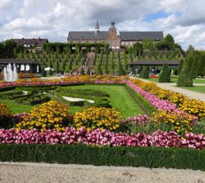 Im Hintergrund das Kloster Kamp, im Vordergrund ein Garten mit leuchtenden pinken und gelben Blumen an einer gepflegten Rasenfläche die von kleinen Buchsbaumhecken umfriedet ist. Die Laufwege sind mit Kies gefüllt.