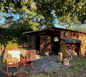 Im Hintergrund stehende Hütte aus Bretter, im Vordergrund stehen zwei rote Schaukelstühle mit Schaffellen. Neben und über der Hütte befinden sich Äste mit grünen Blättern.